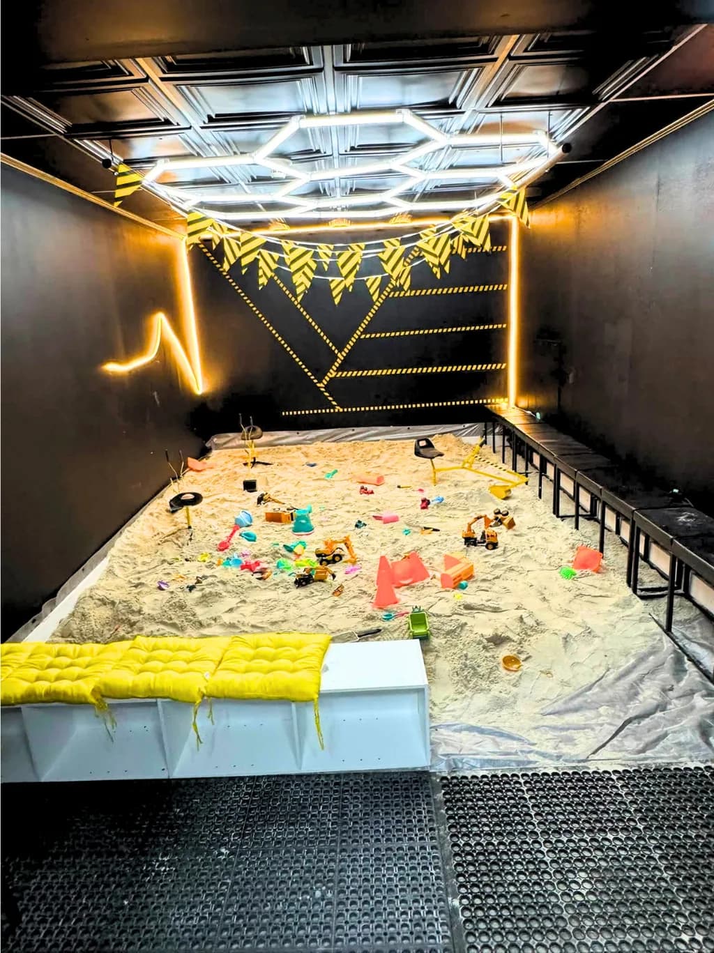 Indoor sand pit with digging toys and construction trucks under yellow caution bunting.